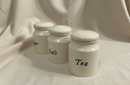 Three white ceramic canisters labeled 'Sugar', 'Salt', and 'Tea' on a beige fabric background.